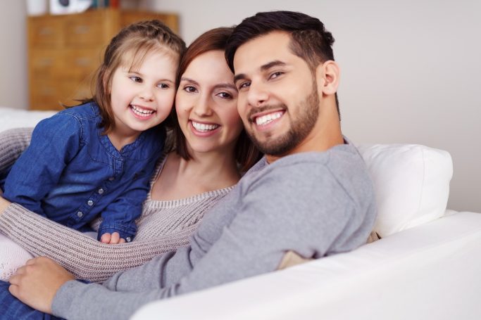 Baufamilie Familie auf einer Couch, lächelnd, mit einem Kind zwischen den Eltern.