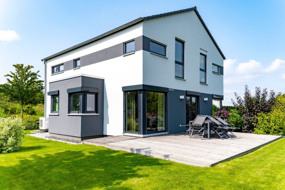 Massivhaus Modernes, zweigeschossiges Haus mit Garten und Holzterrasse unter blauem Himmel.