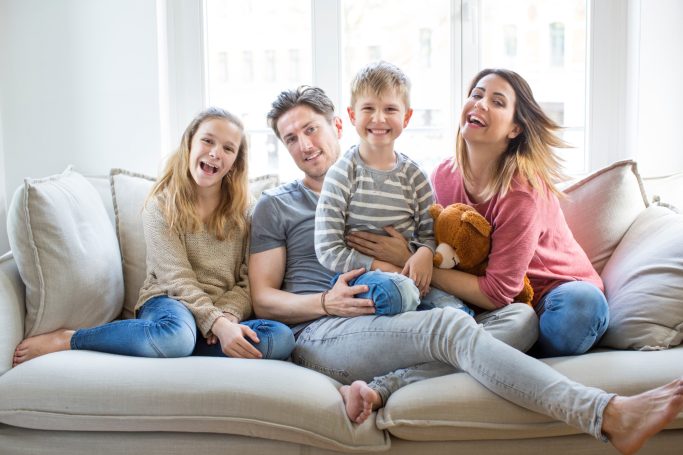 Baufamilie Eine fröhliche Familie sitzt zusammen auf einem Sofa und lächelt in die Kamera.