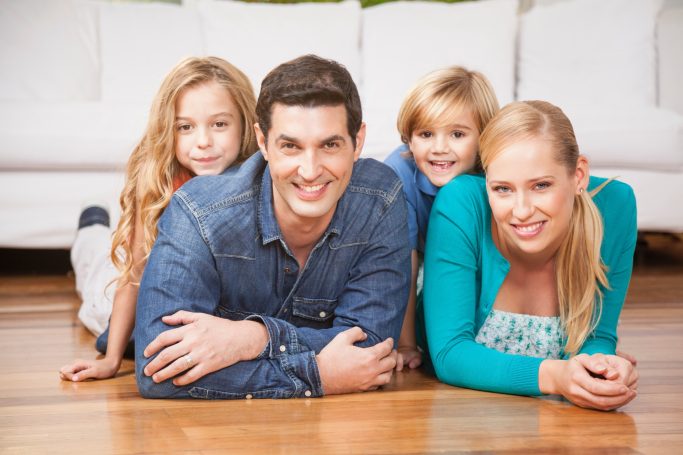 Baufamilie Familie posiert lächelnd im Wohnzimmer auf dem Boden, Kinder liegen vor den Erwachsenen.