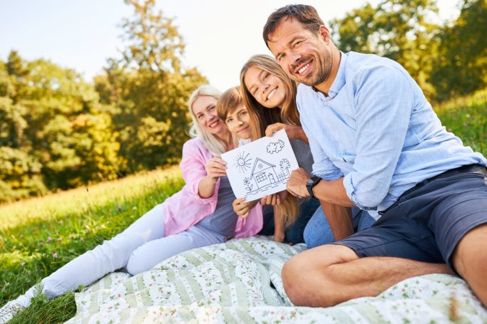 Baufamilie Eine Familie sitzt auf einer Decke im Freien und hält eine Zeichnung hoch.