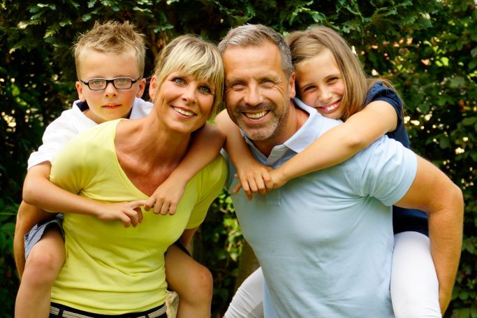 Baufamilie Familie mit zwei Kindern, das Paar lächelt, die Kinder sitzen auf den Schultern der Eltern.